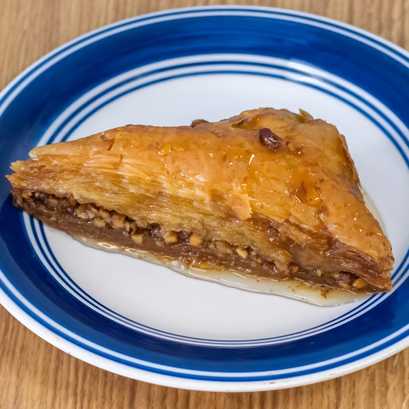 A triangular slice of Greek baklava, glistening with honey syrup and nuts, on a blue and white plate.