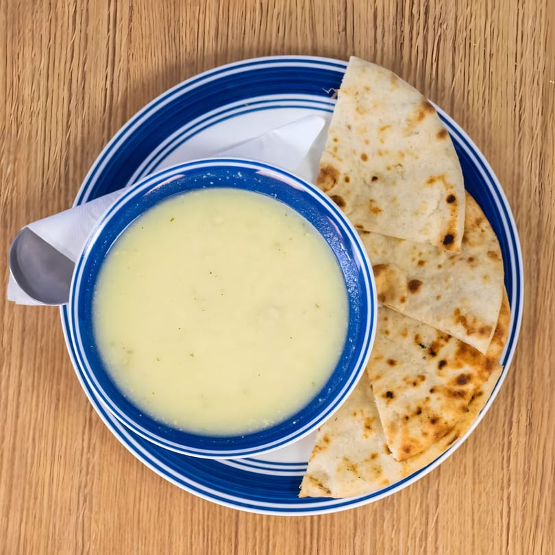 Overhead view of a blue and white bowl of Greek avgolemono soup with pita bread on a wooden table.