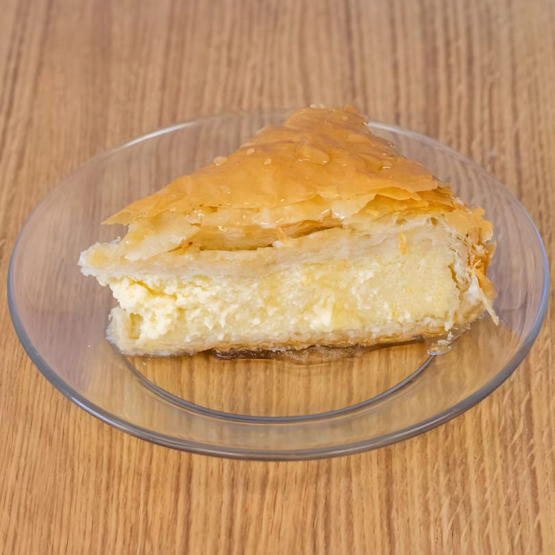 A slice of Greek Gyros' creamy galaktoboureko dessert with flaky pastry and honey on a clear plate.