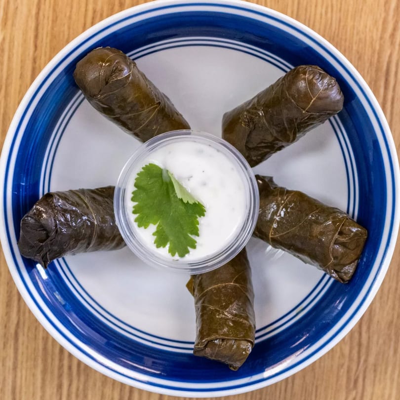 Five Greek dolmades (stuffed grape leaves) arranged around a small cup of tzatziki sauce with cilantro.