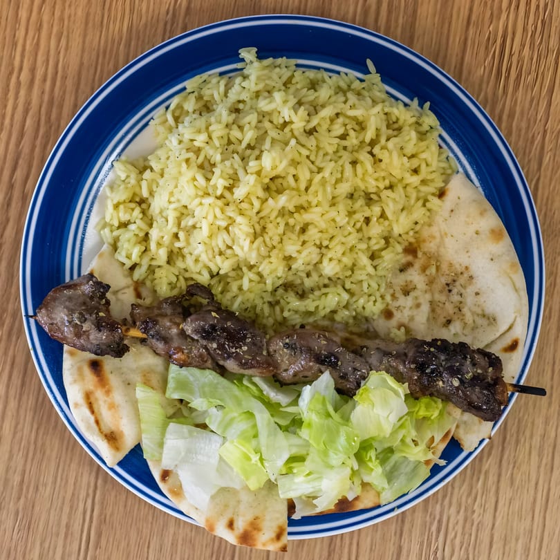 Greek Gyros' plate with a meat skewer, yellow rice, pita bread, and lettuce on a blue-rimmed plate.